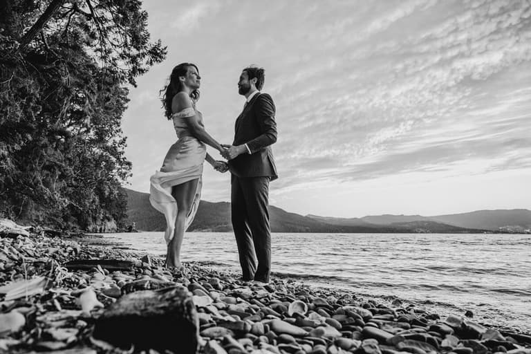 Couple holding hands on a pebble beach, dress catching the wind