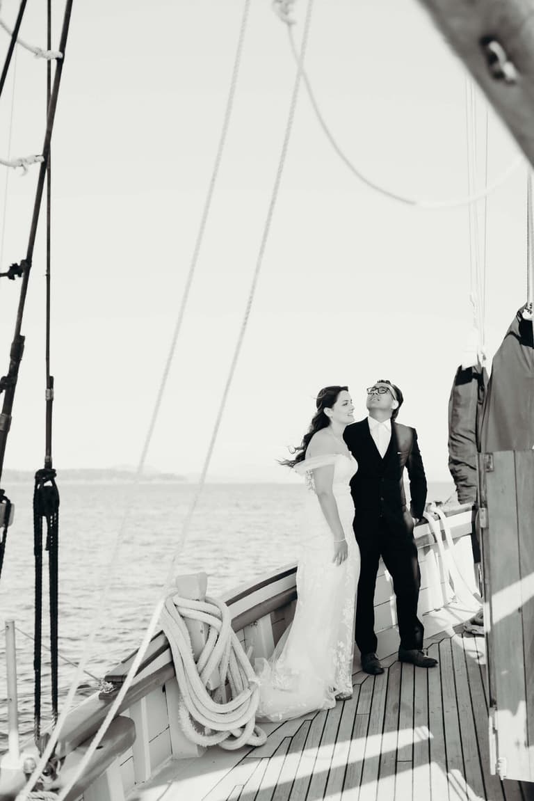 Bride and groom stand on the wooden deck of a sailing ship, tall white canvas sails billowing behind them in black and white