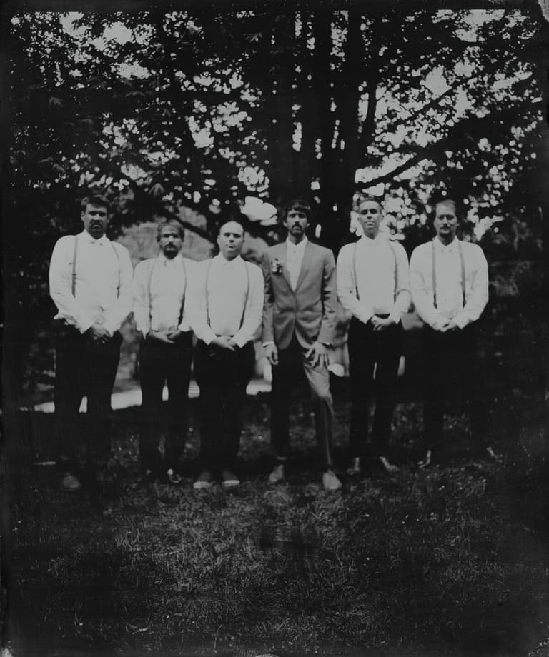 Six men in suspenders and shirtsleeves posing under shade trees