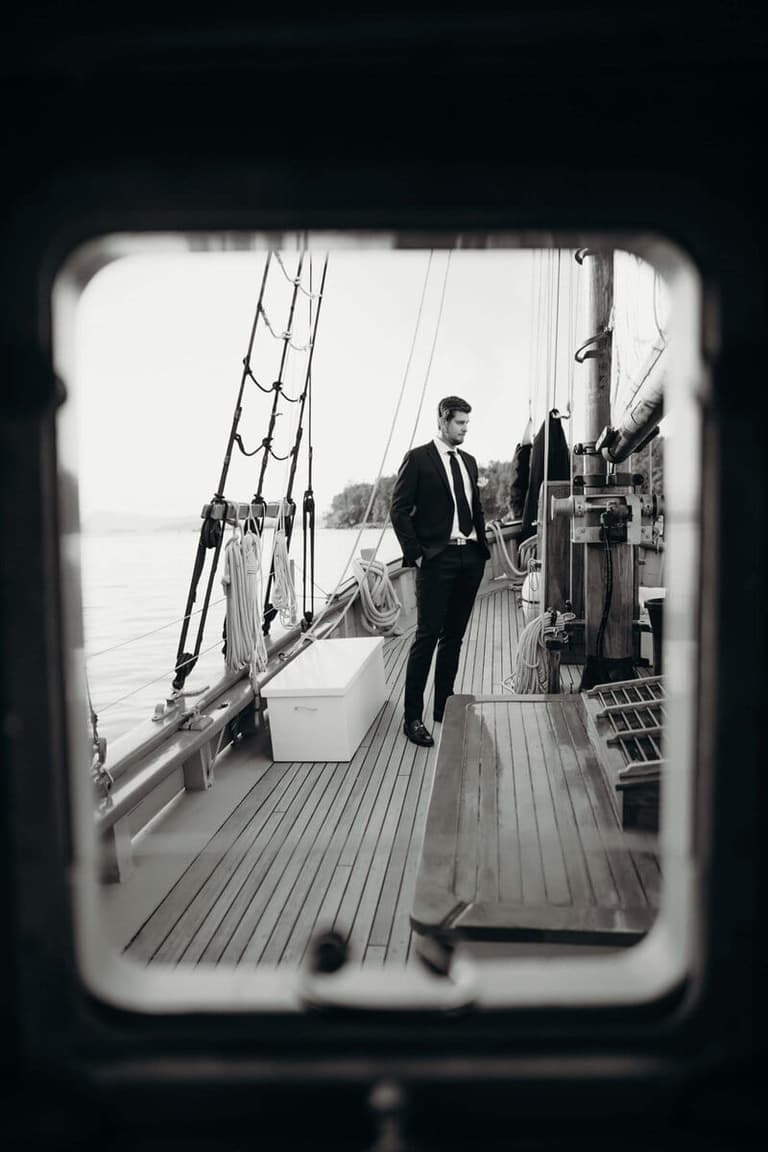 Man in dark suit standing on wooden sailboat deck with rigging and calm water visible beyond