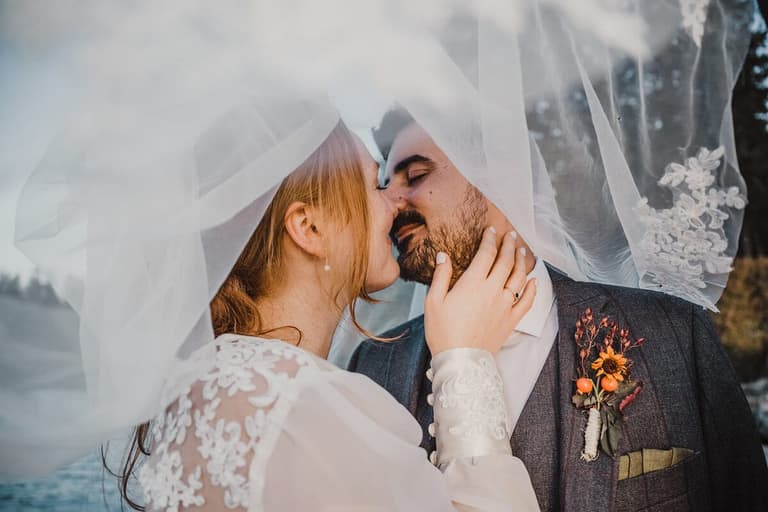 Couple kissing beneath a sheer veil, orange boutonniere visible