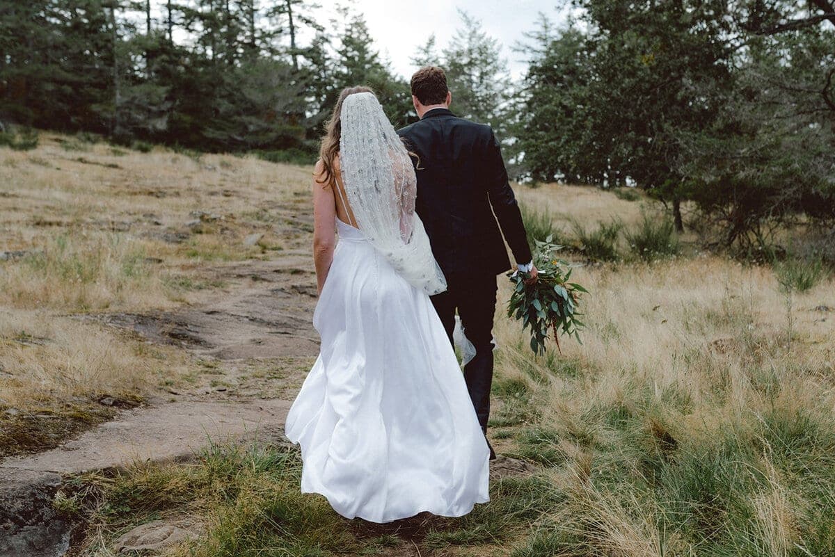Newlyweds climbing a mossy hillside trail with greenery bouquet