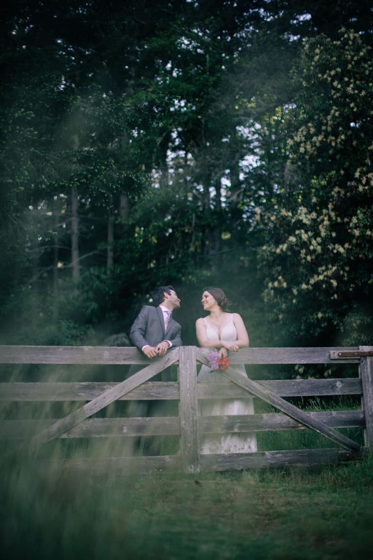 Newlyweds trading glances across the top rail of a gate
