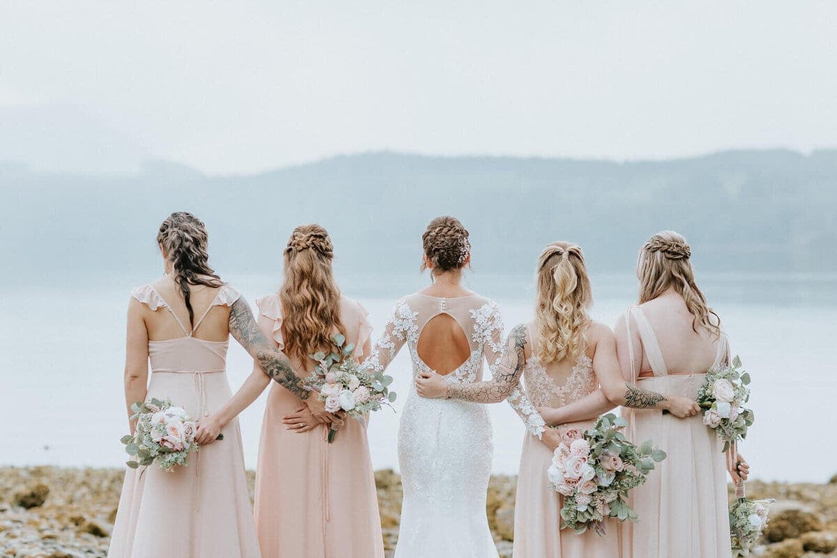 Bride and bridesmaids in blush linking arms on a misty shoreline