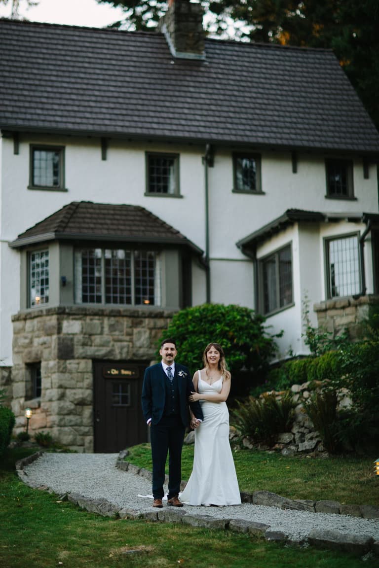 Bride and groom stand arm-in-arm on gravel path before white cottage with dark roof