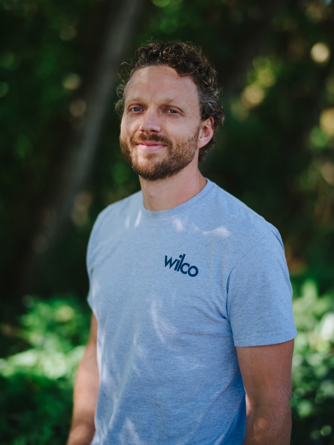 Man with curly brown hair and beard wearing light blue Wilco t-shirt, smiling at camera against blurred green background