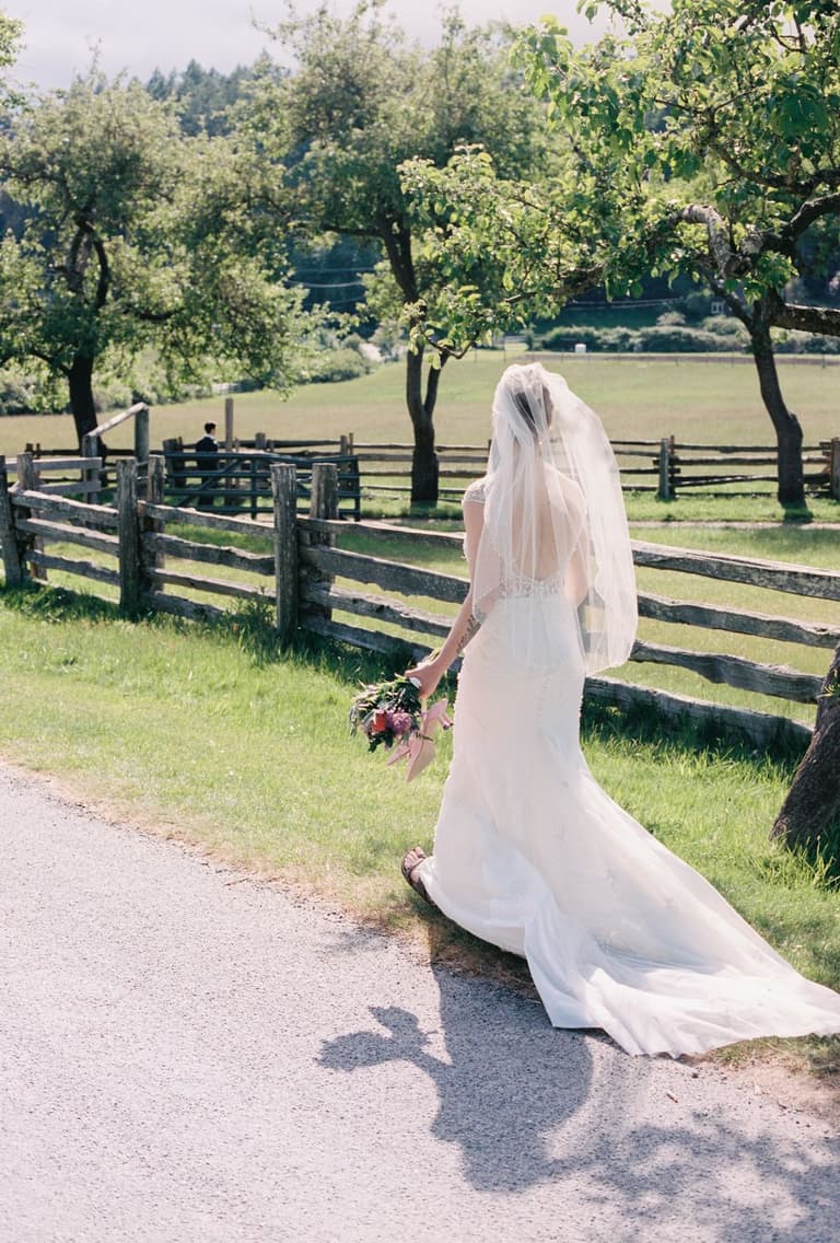 Bride in white embroidered dress holding romantic bouquet of mauve and coral flowers standing barefoot on grass