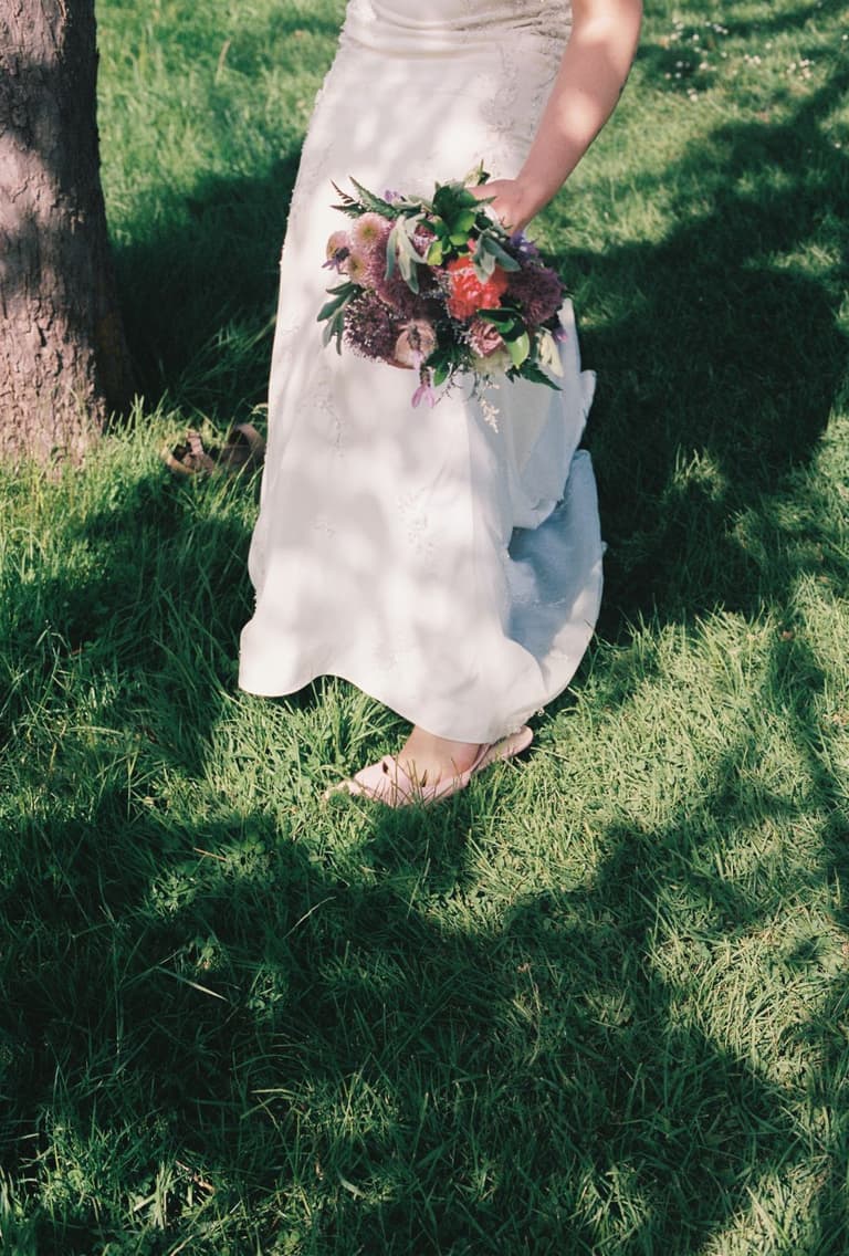 Bride in white gown and veil holds bouquet on gravel path beside wooden fence and blooming fruit trees