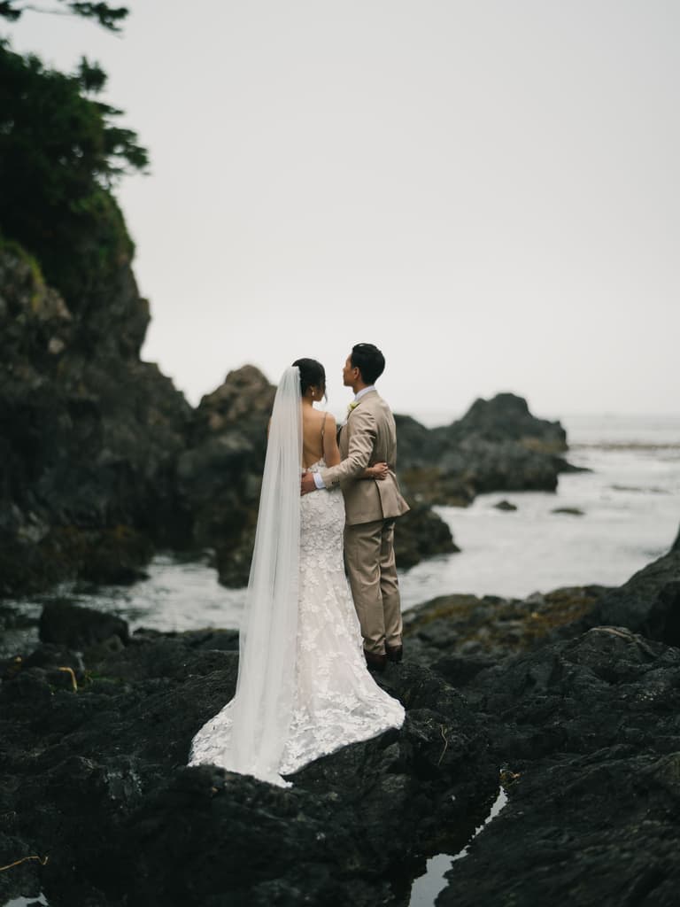 Bride and groom face each other on a rocky outcrop overlooking water, her veil trailing behind them