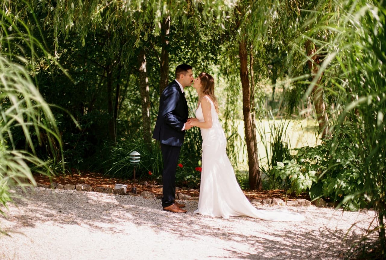 Bride and groom kiss under a natural archway of hanging willow branches, grain and soft light visible on film